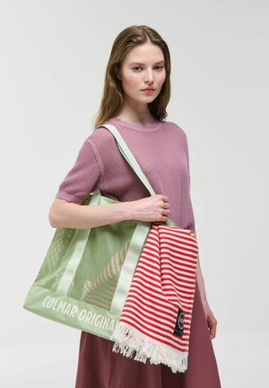 Geantă de plasă verde cu accente albe și logo, care ține o pătură în dungi roșii și albe cu franjuri. Model purtând un top roz tricotat.