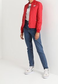 Personne portant une veste matelassée rouge, un t-shirt graphique blanc, un jean bleu et des baskets montantes blanches avec un motif à carreaux noir.