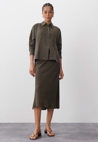 Olive green linen shirt with long sleeves and button closure, paired with a matching midi skirt featuring a side slit. Black heeled sandals.