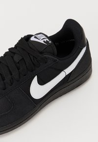 Baskets Nike noires avec logo Swoosh blanc, lacets noirs et col rembourré, photographiées sur un fond clair.