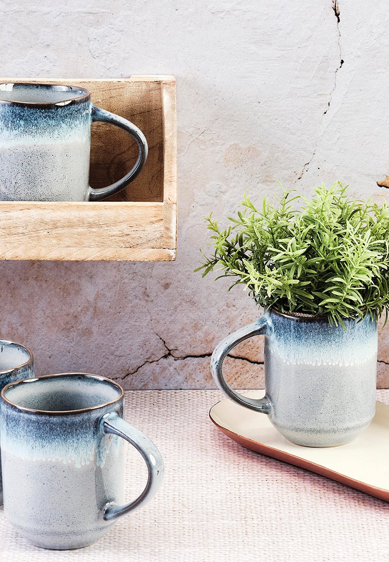 Ceramiczne kubki z gradientowym niebieskim i szarym szkliwem, z okrągłymi uchwytami. Jeden kubek zawiera małą zieloną roślinę, umieszczoną na jasnej tacy.