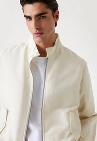 Cream-colored lightweight jacket with a stand collar, front zipper, and side pockets. Features a smooth, textured fabric with minimal stitching.