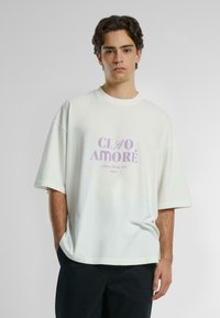 Overdimensjonert hvit bomulls t-skjorte med kort erm som har lys lilla grafisk tekst "CIAO AMORE" sentrert på fronten. Uformell design.