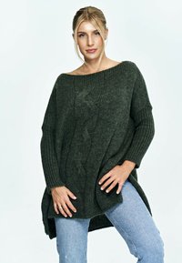 Ciemnozielony, oversize'owy sweter z dzianiny o wytłaczanym wzorze i detalu w splot kablowym. Posiada szeroką, łódkowatą dekoltę oraz szczeliny po bokach, zestawiony z dżinsami.