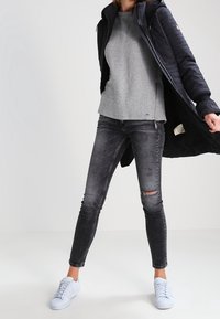 Abrigo acolchado oscuro, suéter gris de canalé, pantalones ajustados negros desgastados y zapatillas de deporte azul claro. Un atuendo casual y en capas con texturas variadas.