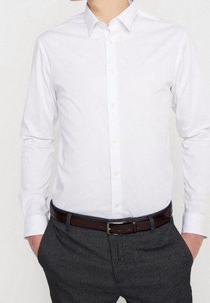 Homme portant une chemise blanche ajustée rentrée dans un pantalon gris foncé avec une ceinture marron foncé, les mains dans les poches.