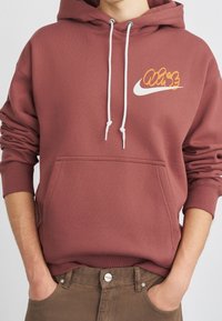 Personne portant un sweat à capuche rose poudré avec des cordons blancs et une poche, associé à un pantalon marron, les mains dans les poches.