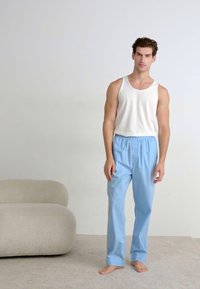 Weißes Baumwoll-Tanktop kombiniert mit hellblauen, locker sitzenden Hosen. Sanfte Textur mit elastischem Bund, stehend auf einem neutral gefärbten Teppich.