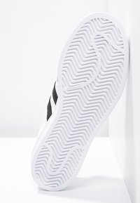 Valkoiset tennarit, joissa on teksturoitu kumipohja, mustilla raidallisilla yksityiskohdilla ja sileällä päällismateriaalilla. Brändäys näkyvissä pohjassa.