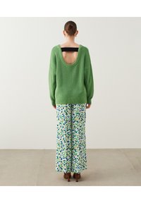 Grüner gestrickter Pullover mit tiefem Rundrücken, kombiniert mit weiten Hosen mit einem bunten abstrakten Muster in Blau, Grün und Weiß.