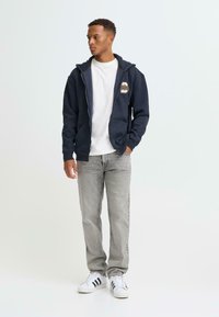 Sweat à capuche zippé bleu marine avec un logo, associé à un t-shirt blanc, un jean gris et des baskets blanches à rayures noires. Tenue décontractée, coupe ample.