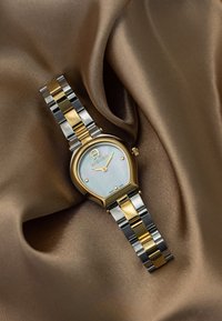 Kultakello ja hopeakello kahden värin yhdistelmä, jossa on pisaran muotoinen kotelo, helmiäismäisestä mussa valmistettu kellotaulu ja kiillotetut metalliset rannekkeet ruskeaa kangasta vasten.