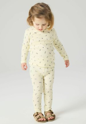 Jeune enfant aux cheveux châtain clair portant un haut à motif floral jaune pâle et des leggings, regardant vers le bas, debout devant un fond uni.