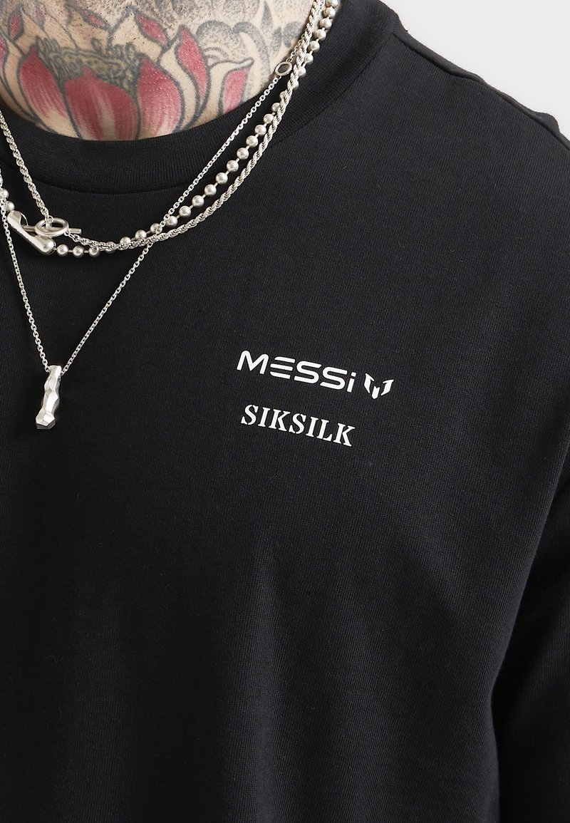 T-shirt en coton noir avec des logos imprimés blancs "MESSI" et "SIKSILK". Des chaînes en argent superposées ajoutent des détails au col.