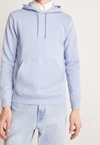 Světle modrá mikina s kapucí a přední kapsou, vyrobená z měkké látky, s malou logovou nášivkou na hrudi a žebrovanými manžetami.