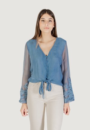 Blusa leggera blu chiaro, trasparente, con maniche lunghe ricamate, scollatura a V, bottoni frontali e dettaglio della vita legato. Abbinata a pantaloni chiari.