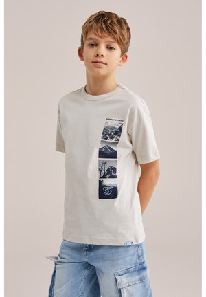 Pois helepruuni juustega, kandes beeži T-särki mägede ja looduse mustritega ning helesiniseid cargo-shorte, seisab käed selja taga.