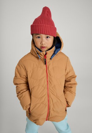 Beige gesteppte Pufferjacke mit blauer Fütterung und rotem Reißverschluss. Kind trägt eine hohe, gerippte rote Strickmütze. Hellblaue Hose.