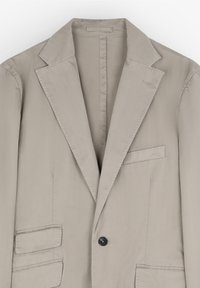 Chaqueta beige con un solo botón negro, solapas con muesca y dos bolsillos frontales. Tejido de textura suave, interior sin forrar.