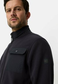 Schwarze Fleecejacke mit hohem Kragen, Reißverschlussverschluss und einer Brusttasche mit Druckknopf. Schwarzes Patch am Ärmel mit Logodetail.