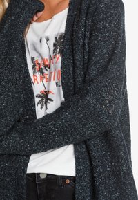 Mørkeblå strik cardigan med en tekstureret overflade, båret over en grafisk hvid t-shirt med palmeprints og koralfarvet skrift.