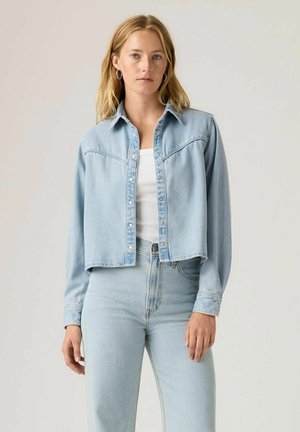 Jasnoniebieska dżinsowa kurtka typu cropped z zatrzaskami, kołnierzykiem z wierzchołkami i długimi rękawami. Zestawiona z dopasowanymi, wysokimi jeansami. Prosty design.