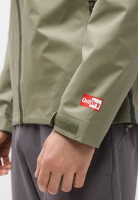 Άτομο που φοράει αδιάβροχο μπουφάν ελιάς με το λογότυπο OutDry στο μανίκι και γκρι παντελόνι, με το χέρι χαλαρό στο πλάι.