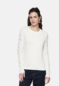 White long-sleeve T-shirt with gathered detail along the sleeves, round neck, and soft texture, paired with dark denim jeans.