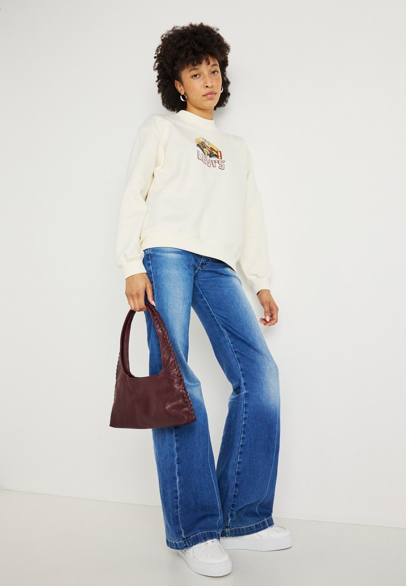 Sudadera crema con gráfico bordado, jeans de campana azules y un bolso de cuero burdeos, combinados con zapatillas blancas.