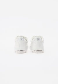 White sneakers with a smooth leather upper, round toe, padded collar, and grey "DKNY" logo on the heel. Textured rubber sole with branding.