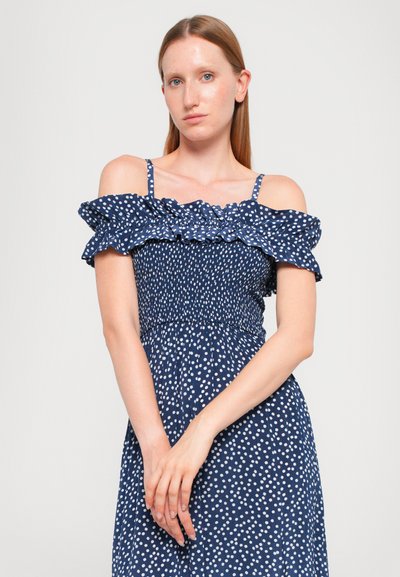 Navy blauwe off-shoulder jurk met witte stippen, gesmokt lijfje en volant halslijn. Spaghettibandjes en vloeiende rok.