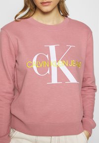 Sudadera de algodón rosa con un logo blanco "CK" y texto amarillo "CALVIN KLEIN JEANS". Puños y dobladillo acanalados, corte relajado.