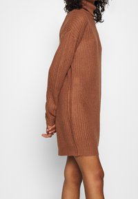 Strick-Turtleneck-Kleid in einem warmen Braunton. Lockere Passform mit langen Ärmeln und geripptem Muster, erreicht die Oberschenkelmitte.