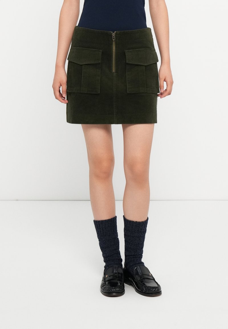 Femme portant une mini-jupe en velours côtelé vert foncé avec une fermeture éclair à l'avant et de grandes poches, associée à des chaussettes foncées et des mocassins noirs.