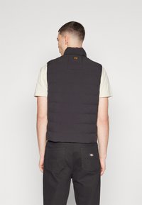 Svart vadderad väst med hög krage, med en liten logotyp baktill. Bärs över en beige skjorta och kombineras med mörka byxor.