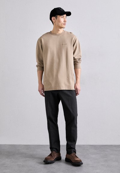 Beige sweatshirt with long sleeves, embroidered logo at chest; paired with dark gray trousers and brown lace-up shoes. Black cap worn.