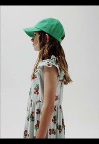 Mladé dievča v bezrukávových šatách s potlačou čerešní a zelenou šiltovkou, stojace bočne proti jednoliatej svetlej stene.