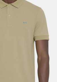 Polo beige con una texture morbida, maniche corte e una pattina con tre bottoni. Presenta un logo ricamato blu sul lato sinistro del petto.