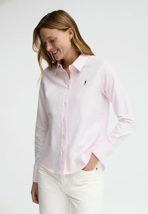 Mujer sonriente con camisa de botones rosa claro con el logo de un jugador de polo en negro y pantalones blancos, con la mano en el bolsillo, sobre un fondo claro y sencillo.