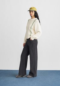 Krem džemper s detaljima zelenog šava, upareno s tamnosivim širokim hlačama i sivim sportskim cipelama, upotpunjeno žutom kapom.