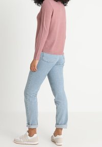 Donna con jeans chiari con risvolto, maglia rosa a maniche lunghe e sneakers bianche, in piedi di profilo contro uno sfondo semplice.
