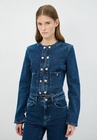 Chaqueta de mezclilla azul oscuro con mangas largas, cuello redondo, botones dorados y dos bolsillos en el pecho. Textura suave con detalles de costura mínimos.