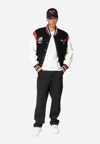 Black and white varsity jacket with red accents, embroidered logos; paired with a white shirt, black pants, and a black cap featuring a logo.
