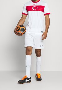 White football kit with a red stripe featuring the Turkish flag; paired with black and orange cleats, holding a black and orange soccer ball.