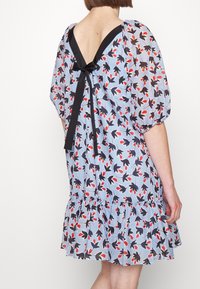 Tył niebieskiej, kwiatowej sukienki do kolan z bufkami na rękawach, falbaną na dole, głębokim dekoltem w kształcie litery V na plecach oraz czarną kokardą jako ozdobą.