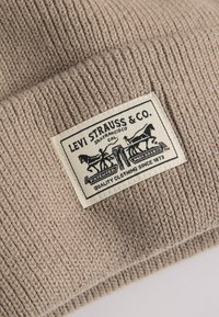Beige gebreide muts van textuurstof. Bevat een rechthoekig stoffen label met een zwart logo, tekst en afgebeelde paarden.