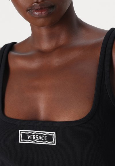 Μαύρη ριγέ αμάνικη μπλούζα με τετράγωνη λαιμόκοψη και λευκή κεντητή ετικέτα "VERSACE" στο μπροστινό μέρος, που αναδεικνύει την υφή και τον σχεδιασμό.