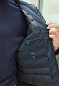 Jachetă puf albastră marină cu un exterior neted și căptușeală matlasată, dotată cu o pungă interioară și eticheta "NEXT". Închidere cu fermoar vizibilă.