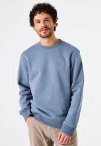 Sudadera azul con cuello redondo y puños acanalados, que presenta un pequeño logo bordado en el pecho. Tejido suave, corte relajado.