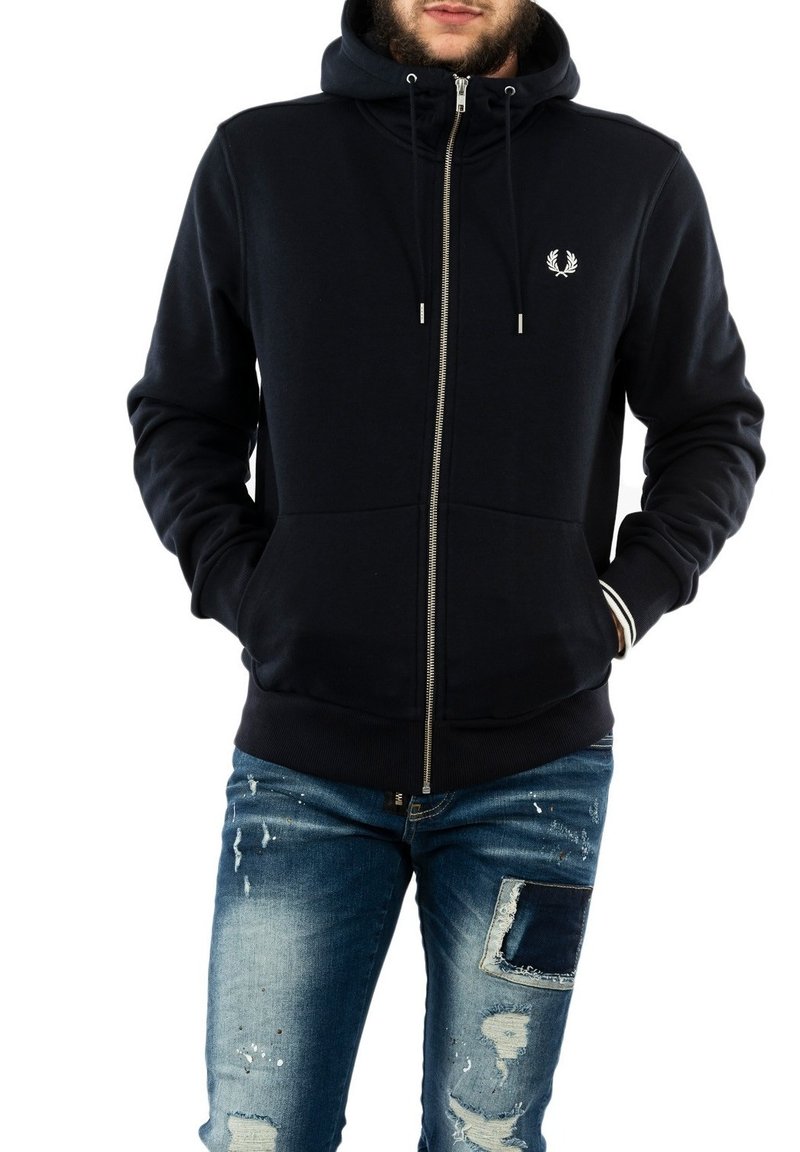 Felpa zipata blu navy con tasche frontali, cappuccio con cordino, polsini a costine e dettaglio del logo sul petto; abbinata a jeans in denim strappati.